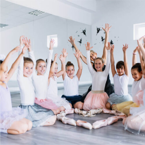 Ballet kids sitting on the ground, reaching their hands in the air, with their shoes on the ground making the shape of a heart