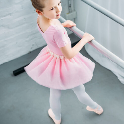 A young white ballet dancer wearing a pink tutu, practicing with her hands on the bar