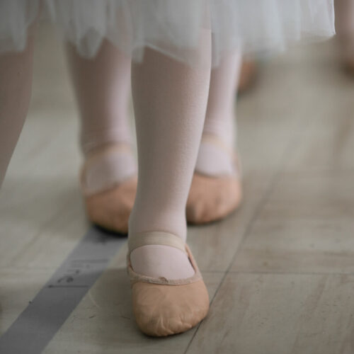 closeup of toddler ballet dancer shoes, with white tights, standing on a wood floor