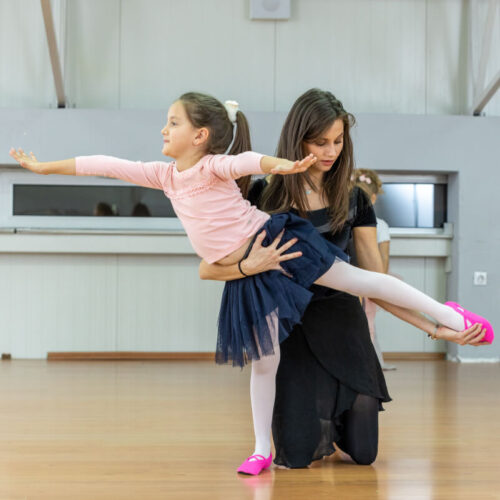 A ballet teacher helping a little girl pose with one foot off the ground and her arms outstretched, wearing a navy tutu and pink ballet shoes in a dance studio
